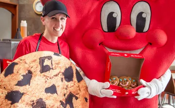 A YMCA mascot is holding an open box of smile cookies and posing beside a young female Tim Hortons Store Owner who is dressed as a large cookie. 