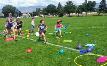 A female YMCA camp leader runs through an outdoor obstacle course with 6 children, aged 7-9, outside on a sunny day. 
