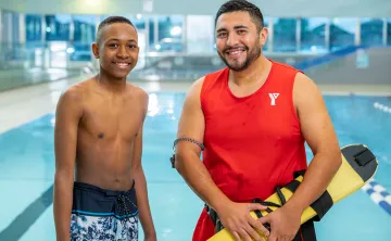 A young teen in swim trucks stands beside a male lifeguard with dark hair in front of a pool. They are smiling at the camera. 