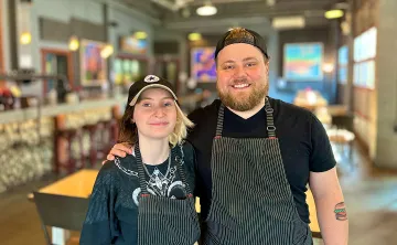 A young man and woman in black striped chef aprons are standing in a restaurant with their arms around one another and smiling at the camera. 