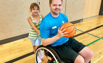 A young blonde man in a blue shirt and a wheelchair is smiling at the camera and holding a basketball in a gymnasium. There's an 8 year old girl holding the back handles of the chair also smiling at the camera. 
