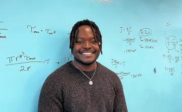 A young man with short black thin dreadlocks in a brown sweater is smiling at the camera in front of a teal board with mathematical equations scribbled on it.