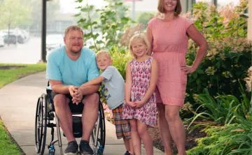 A family of 4 with two young children under the age of 7 are standing together outside in the sunshine and smiling at the camera.