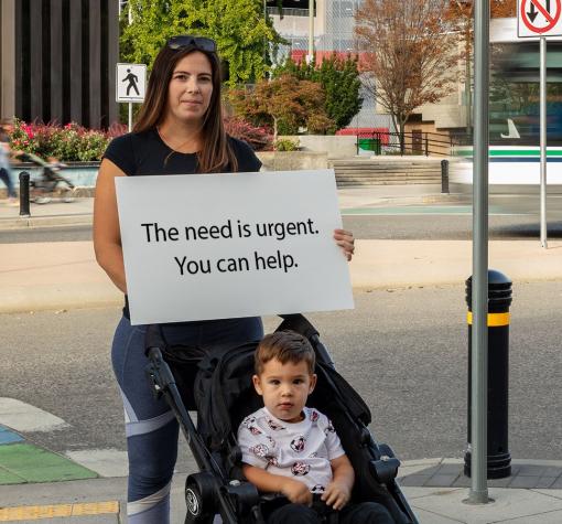 Y Give (teaser) Mother holding sign that says: The need is urgent. You can help.