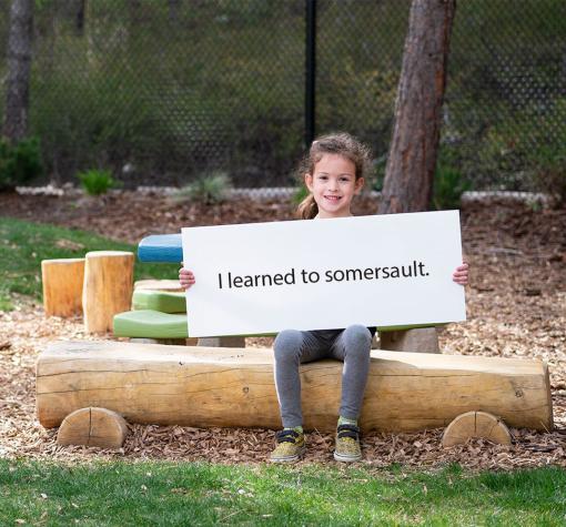 AR 2021 FA Teaser 6 year old girl sitting on a bench smiling and holding a sign that reads 'I learned to somersault.' There are trees and a yellow slide in the distance.